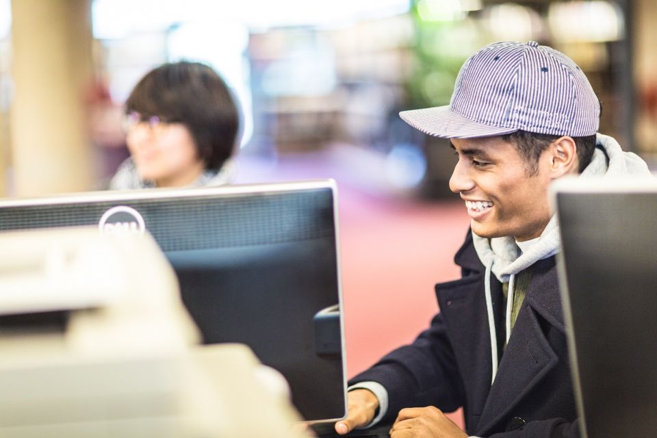 Jongen met pet op zit in de bibliotheek achter een computer, lachend.